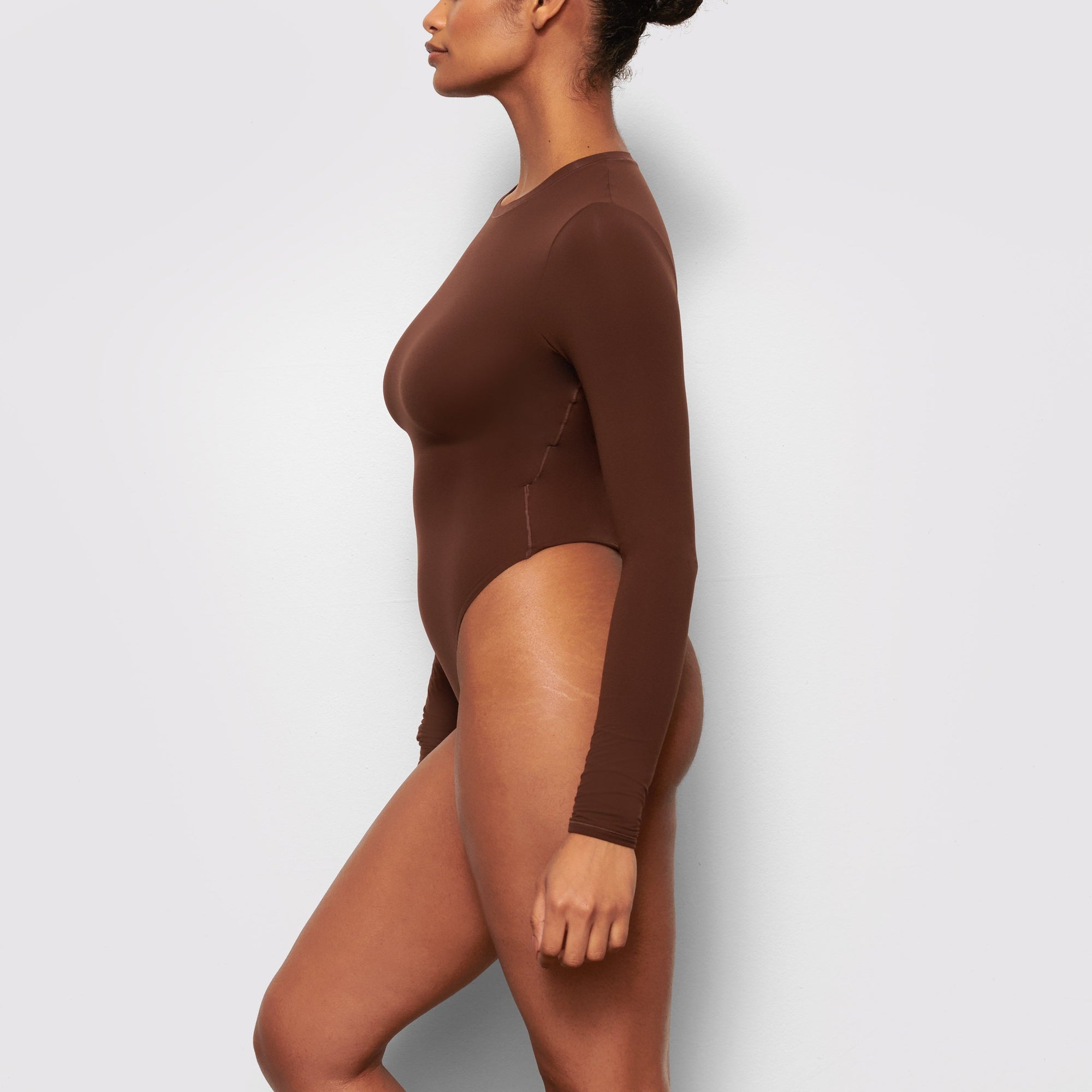A MODEL STANDS SIDEWAYS SHOWING THE PROFILE OF THE FITS EVERYBODY LONG SLEEVE CREW NECK BODYSUIT | COCOA